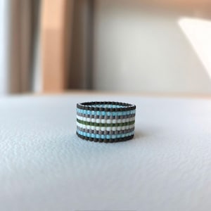 Puede incluir: Un anillo de cuentas con un patrón de rayas. El anillo presenta bandas alternas de cuentas negras, azules, grises, blancas y verdes. El anillo mide aproximadamente 2 cm de altura y 2,5 cm de diámetro.