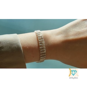 Puede incluir: Una pulsera plateada con un diseño de eslabones rectangulares se muestra en una muñeca. La pulsera está hecha de pequeñas piezas rectangulares conectadas entre sí. El fondo es de color azul claro.