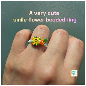 May include: A beaded ring featuring a yellow smiley face flower with green, orange, and white beads. The text A very cute smile flower beaded ring is visible above the ring. A small logo with a diamond icon and the text DIYbyRim is in the corner.