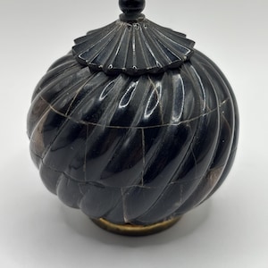 May include: A round, black, lidded container with a spiraling, ribbed texture. The lid features a small, spherical knob at the top. The body is constructed from segmented pieces with visible seams, and the base is finished with a metallic gold ring.