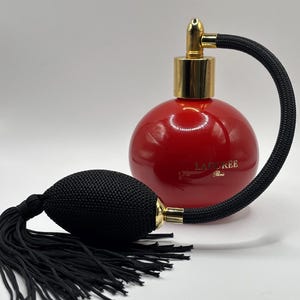 May include: A round, glossy red perfume bottle with a gold-toned metal atomizer nozzle. A black fabric-covered hose connects the nozzle to a black, oval-shaped bulb pump featuring a long, decorative black tassel. The text LADURÉE Paris is printed in gold.