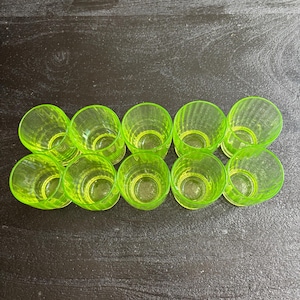 Op de afbeelding: Een set van tien kleine, doorschijnende, neongeel-groene drinkglazen gerangschikt in twee rijen van vijf. Elk glas heeft een cilindrische vorm met verticale ribbels langs de buitenwanden en een gladde, cirkelvormige rand aan de bovenkant.