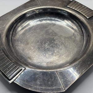 Puede incluir: Una bandeja cuadrada de metal plateado con un centro circular hundido. El borde ancho y plano presenta detalles geométricos, con dos lados opuestos que muestran una serie de ranuras verticales paralelas, mientras que los otros dos lados son lisos.