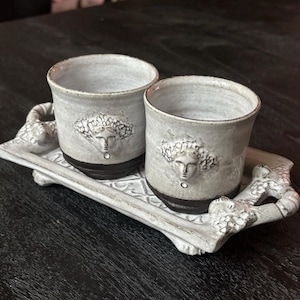 Può includere: Due tazze in ceramica con una sezione superiore smaltata bianca e una base marrone scuro, ciascuna caratterizzata da un rilievo di un volto. Le tazze poggiano su un vassoio rettangolare in ceramica abbinato con manici e superficie lavorata.