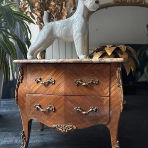 May include: A small wooden chest of drawers with two drawers, featuring a light brown wood veneer in a chevron pattern, gold-toned metal handles, curved legs with gold-toned accents, and a marble top, topped with a white ceramic dog figurine.