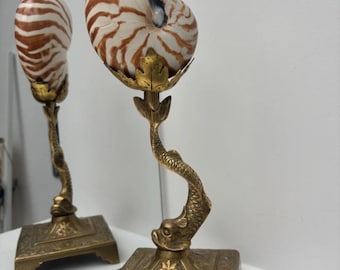 May include: A nautilus shell with brown and white stripes mounted on a gold-toned metal stand. The stand features a square base with ornate engravings, a central column shaped like a fish, and a top holder designed as a stylized leaf structure.