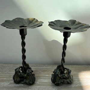 May include: A pair of dark metal candle holders featuring a twisted stem design. Each holder has a wide, flat, petal-shaped top and a base formed by three sculpted frogs arranged in a circle, supporting the central vertical twisted column.