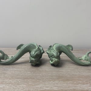 Könnte beinhalten: Ein Paar salbeigrüne, dekorative Delfinfiguren mit strukturierten Schuppen und geringelten Schwänzen. Jeder Delfin ist in einer geschwungenen, horizontalen Position mit detaillierten Gesichtszügen, Flossen und einer matten Oberfläche geformt.