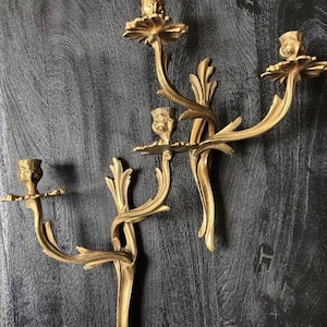 May include: A pair of gold-colored metal wall sconces, each featuring two candle holders. The sconces have a branching, organic design with leaf-like details and curved arms that extend outward to support the candle cups at different heights.