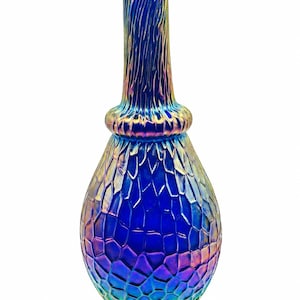 Op de afbeelding: Een hoge, iriserende blauwe glazen vaas met een bolvormige basis met een textuur van schubben en een slanke, verticaal geribbelde hals. Het oppervlak vertoont een metallic, veelkleurige glans die verschuift tussen diepblauwe, paarse en gouden tinten.