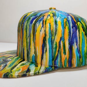 May include: A baseball cap with a flat peak, featuring a striking, abstract design. The cap is adorned with a flowing pattern of paint in shades of blue, yellow, orange, and green. The peak also has the same colourful design.