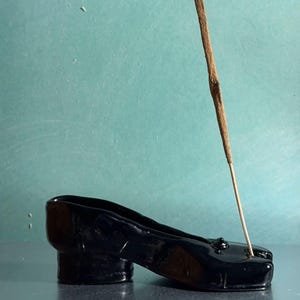 Könnte beinhalten: Ein schwarzer Keramik-Schuh-förmiger Räucherstäbchenhalter mit kleinem Absatz. Ein Räucherstäbchen ist in den Schuh gesteckt. Der Hintergrund ist türkis.