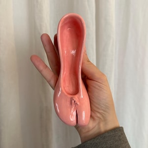 May include: A pink ceramic spoon rest shaped like a ballet slipper. The spoon rest has a glossy finish and a split toe design. The object is held in a hand against a white curtain background.
