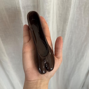 Könnte beinhalten: Ein dunkelbrauner Schuh mit geteilter Zehenpartie, der einem Ballettschuh ähnelt, wird in einer Hand gehalten. Der Schuh hat eine glänzende Oberfläche und ein kleines dekoratives Detail an der Spitze. Der Hintergrund ist ein weißer Vorhang.