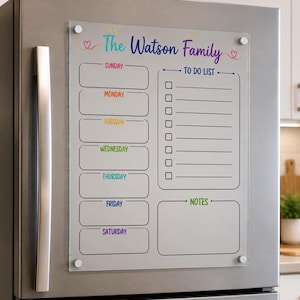 May include: A clear acrylic magnetic refrigerator planner featuring the text The Watson Family at the top. It includes a weekly schedule with sections for Sunday through Saturday, a vertical to-do list with checkboxes, and a notes section at the bottom.