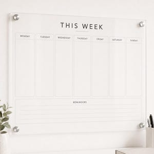 May include: A clear acrylic weekly wall planner featuring the text THIS WEEK at the top, with seven vertical columns labeled Monday through Sunday, and a section at the bottom labeled REMINDERS with four horizontal lines, secured by silver hardware.