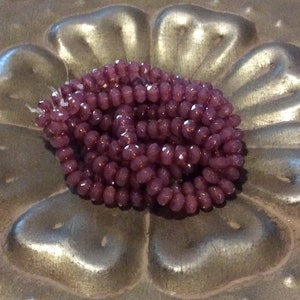 May include: A strand of small, faceted glass beads in a deep purple color. The beads are arranged in a coil shape and are displayed on a silver background.