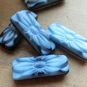 May include: A group of light blue glass flower-shaped beads with black accents. The beads are rectangular in shape and have a raised flower design.