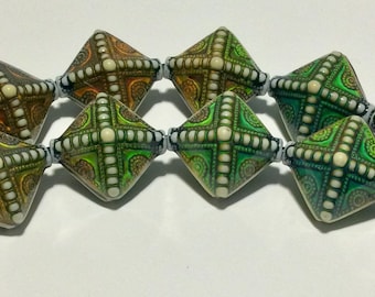 May include: A strand of colorful, patterned, diamond-shaped beads. The beads are made of polymer clay and have a variety of colors, including green, brown, and white.