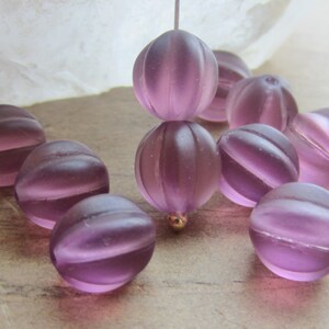 May include: A collection of frosted, lavender-colored glass beads. The beads have a ribbed, spherical design with a subtle, striped effect. Some beads are strung together, and a few are scattered.