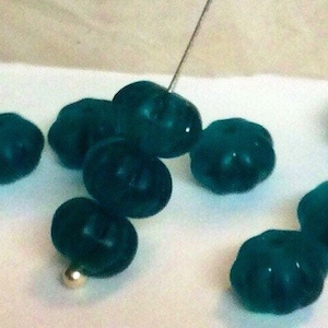 May include: A collection of teal blue glass beads with a textured surface. The beads are round and have a slightly flattened shape.