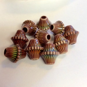 May include: A group of ten ceramic beads with a brown, white, and green glaze. The beads are shaped like a teardrop with a rounded top and a pointed bottom. The beads have a textured surface with raised lines and dots.