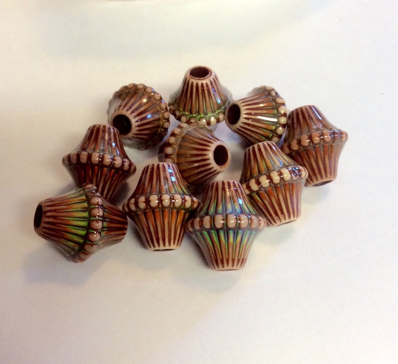 May include: A group of ten ceramic beads with a brown, white, and green glaze. The beads are shaped like a teardrop with a rounded top and a pointed bottom. The beads have a textured surface with raised lines and dots.