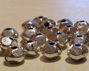 May include: A group of silver metal beads with a faceted design. The beads are arranged in a loose pile on a white surface.