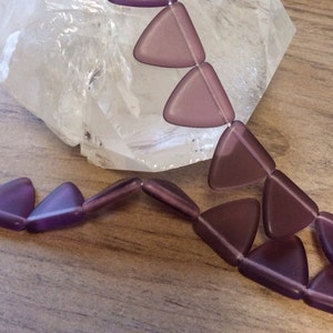 May include: A strand of triangular, matte purple beads. The beads are strung together, creating a necklace or bracelet. The background includes a clear, crystalline rock and a wooden surface.