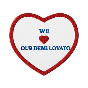 Puede incluir: Parche bordado en forma de corazón con borde rojo y centro blanco. El parche presenta el texto "WE", un corazón rojo y "OUR DEMI LOVATO" en azul. El parche es probablemente para ropa o accesorios.