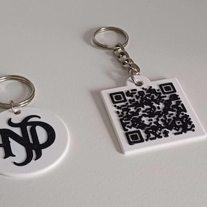 Peut inclure: Deux porte-clés blancs avec des anneaux argentés. L'un est rond avec les initiales noires "ND". L'autre est carré avec un code QR noir. Les deux sont sur une surface blanche.