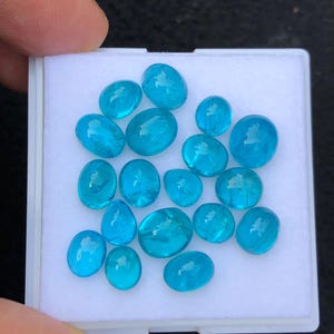 May include: A selection of polished, oval-shaped, blue gemstones of various sizes. The gems are presented in a white, rectangular container, with a hand partially visible. The stones have a translucent appearance.