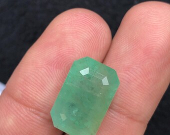 May include: A faceted, rectangular emerald gemstone. The gem is a vibrant green colour with visible internal inclusions. The cut features multiple facets, reflecting light. The emerald is held between fingers, showcasing its size and clarity.