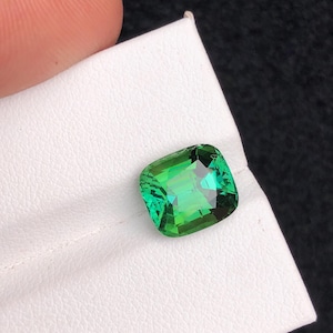 May include: A vivid, emerald-green gemstone with a cushion cut. The gem is held on a white surface, with a dark background. The gemstone's facets reflect light, showcasing its clarity and colour. This gemstone is ideal for jewellery making.
