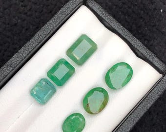 May include: A collection of six loose green gemstones displayed in a black presentation box with a white interior. The set includes three rectangular emerald-cut stones and three oval-cut stones, all featuring faceted surfaces and varying shades of green.