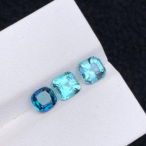 Puede incluir: Tres gemas talladas cuadradas en diferentes tonos de azul, desde el azul verdoso intenso hasta el aguamarina claro, están dispuestas sobre una superficie blanca. Las gemas están facetadas, reflejando la luz y mostrando su claridad.