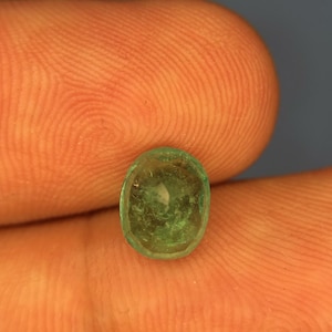 May include: A small, oval-shaped emerald gemstone with a translucent, vibrant green hue and visible internal inclusions. The stone features a faceted surface that catches the light, displaying a natural, slightly irregular cut and a polished finish.