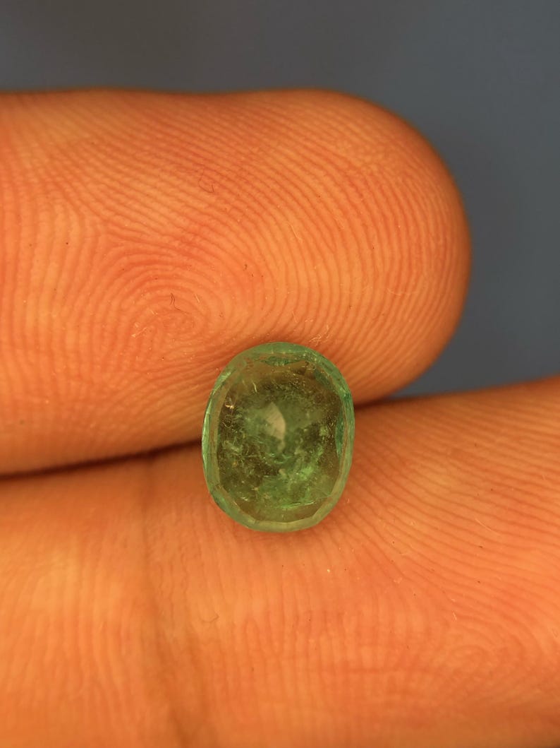 May include: A small, oval-shaped emerald gemstone with a translucent, vibrant green hue and visible internal inclusions. The stone features a faceted surface that catches the light, displaying a natural, slightly irregular cut and a polished finish.