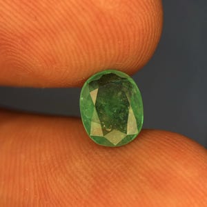 May include: A faceted oval-shaped emerald gemstone with a vibrant green colour and visible internal inclusions. The stone features a polished surface with multiple geometric facets that reflect light, showcasing a deep, saturated green hue throughout.
