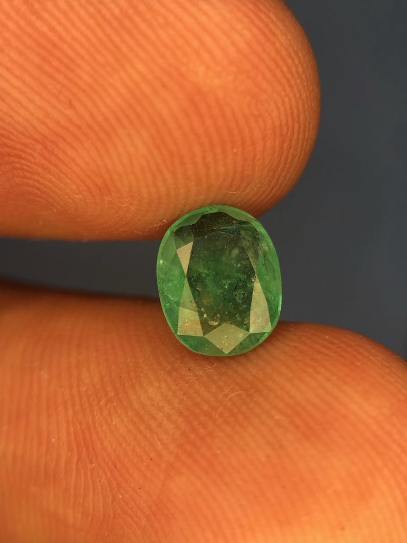 May include: A faceted oval-shaped emerald gemstone with a vibrant green colour and visible internal inclusions. The stone features a polished surface with multiple geometric facets that reflect light, showcasing a deep, saturated green hue throughout.