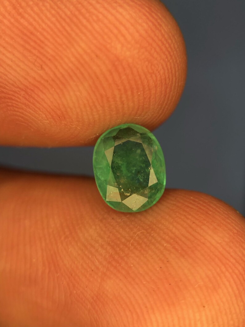 May include: A loose oval-cut gemstone with a vibrant green colour and a faceted surface. The stone displays internal inclusions and a polished finish, reflecting light across its multiple geometric facets. The gem is held between two fingers.