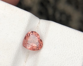 May include: A heart-shaped gemstone with a faceted surface and a light peach-orange colour. The stone is positioned on a white textured surface, displaying clear internal reflections and a polished finish across its multiple cut edges.