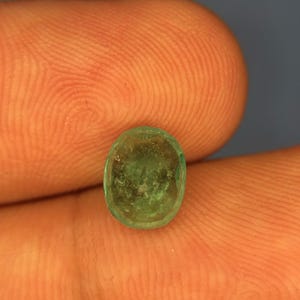 May include: A small, oval-shaped emerald gemstone with a translucent, light green hue and visible internal inclusions. The stone features a faceted surface that catches the light, displaying a natural, slightly cloudy texture throughout its structure.