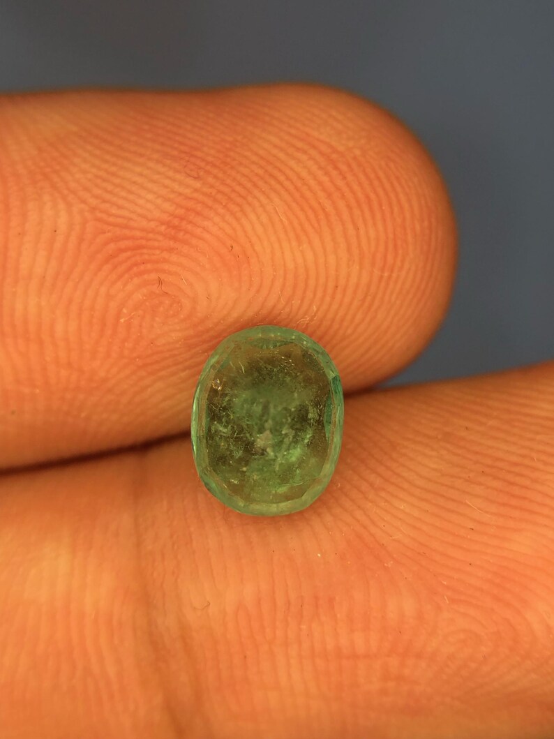 May include: A small, oval-shaped emerald gemstone with a translucent, light green hue and visible internal inclusions. The stone features a faceted surface that catches the light, displaying a natural, slightly cloudy texture throughout its structure.