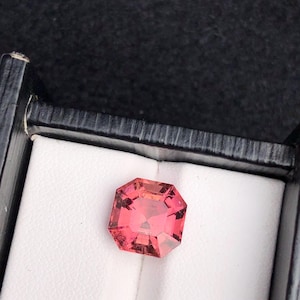 Könnte beinhalten: Ein facettierter, rot-rosa Edelstein, wahrscheinlich ein Turmalin, wird in einer weiß gefütterten Schachtel präsentiert. Der Stein hat eine rechteckige Form mit geschnittenen Ecken, die das Licht reflektieren und seine Klarheit und Farbe zeigen.