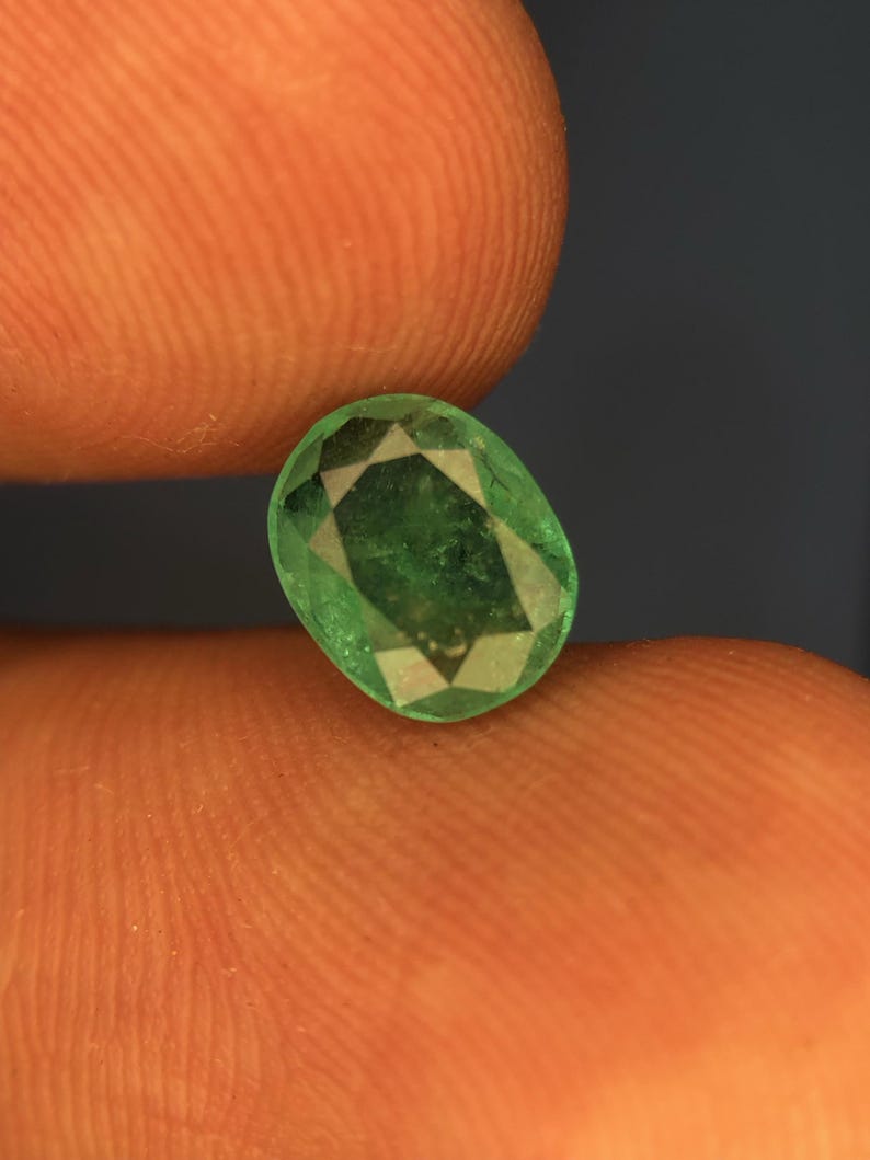 May include: An oval-cut, vibrant green gemstone featuring a faceted surface that reflects light. The stone displays a translucent quality with visible internal inclusions and a polished finish, showcasing a classic, symmetrical geometric cut.