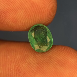 May include: A loose oval-cut gemstone with a vibrant green colour and multiple facets across its surface. The stone displays natural internal inclusions and a polished finish, reflecting light through its transparent structure in a clear, detailed view.