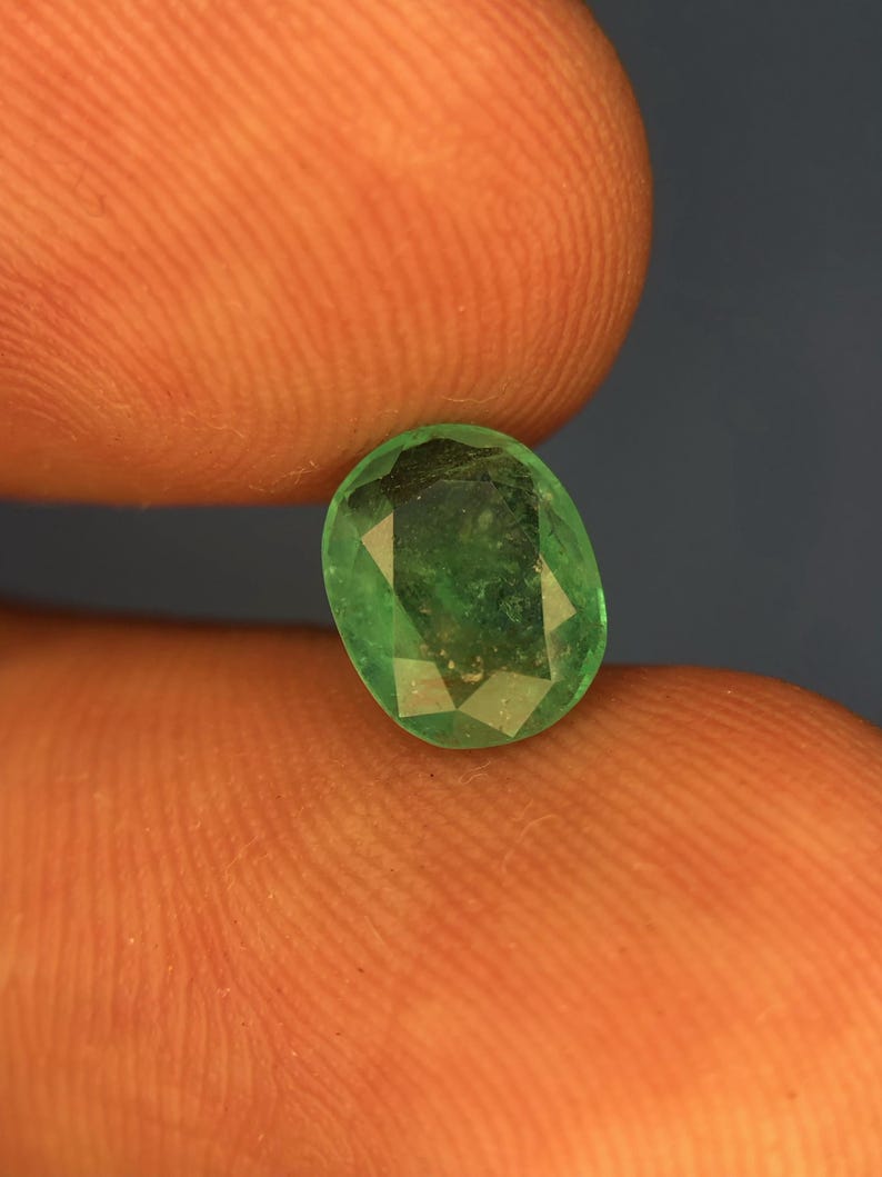May include: A loose oval-cut gemstone with a vibrant green colour and multiple facets across its surface. The stone displays natural internal inclusions and a polished finish, reflecting light through its transparent structure in a clear, detailed view.