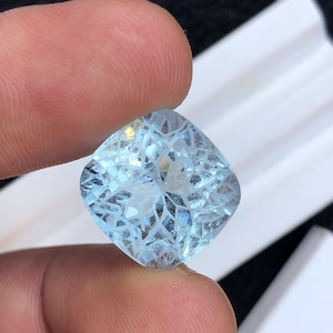 May include: A light blue, cushion-cut gemstone with intricate facets. The gem is held between two fingers, showcasing its clarity and brilliance. The stone's color is a pale, icy blue, reflecting light throughout its many cuts.