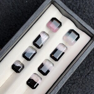 May include: A selection of rectangular gemstones in a black box. The gems feature a black border with a gradient of colours, including pink, grey, and clear. The box has a white interior and a dark grey exterior.